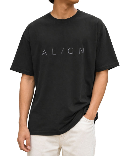 Young male model wearing a black oversized T-shirt with a minimalist “AL/GN” typography design across the chest.