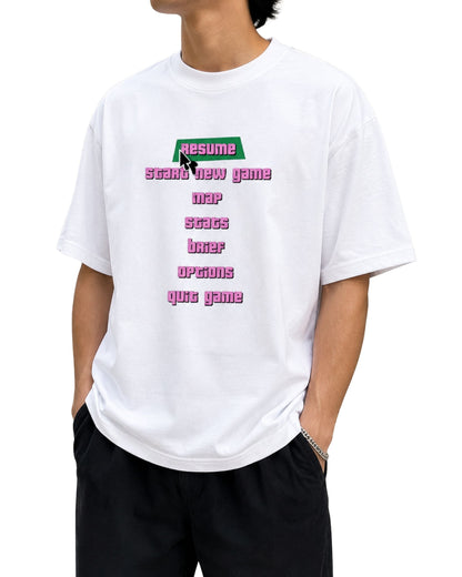Young male model wearing a white graphic T-shirt outdoors, featuring a retro video game menu design with “Resume,” “Start New Game,” “Map,” “Stats,” “Brief,” “Options,” and “Quit Game” in pixel-style typography.