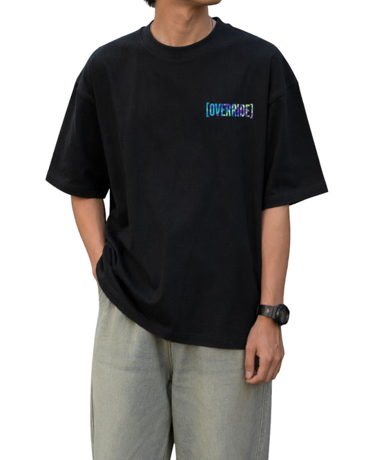 Black oversized T-shirt with a small multicolor glitch-style “OVERRIDE” graphic printed on the left chest.