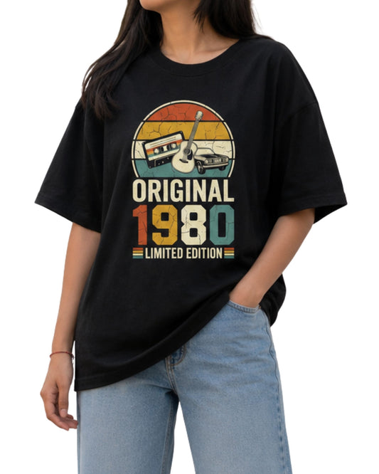 Young female model wearing a oversized graphic T-shirt with a vintage “Original 1980 Limited Edition” design featuring a cassette tape, acoustic guitar, retro car, and distressed sunset colors Black
