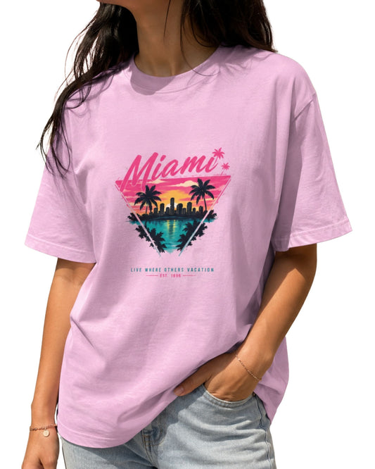 A unisex crew tee featuring a vibrant retro graphic of a Miami sunset inside a triangle, complete with palm trees, a city skyline over water, and the text "Miami" and "LIVE WHERE OTHERS VACATION EST. 1896" light baby pink