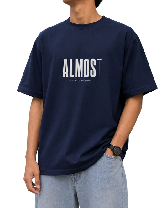 Royal blue oversized T-shirt with a minimalist “ALMOST” typography graphic, finished with a thin stylized “T” and the small tagline “NOT QUITE, BUT CLOSE.” underneath.