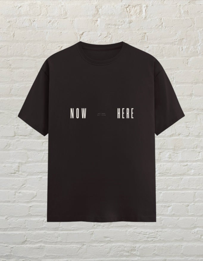 CoreTeez coffee brown unisex cotton t-shirt featuring a minimalist white Now Here graphic print against a brick wall.