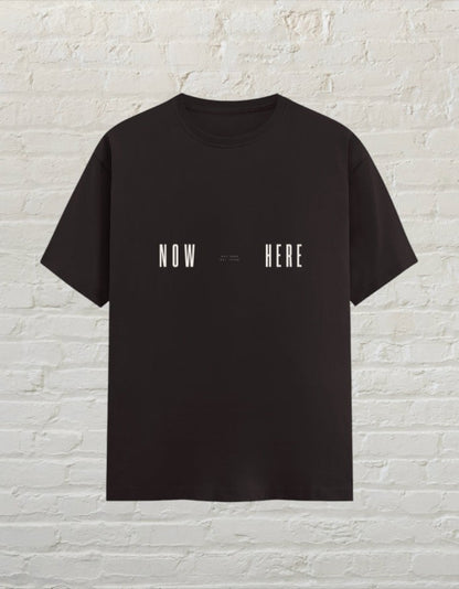 CoreTeez coffee brown unisex cotton t-shirt featuring a minimalist white Now Here graphic print against a brick wall.