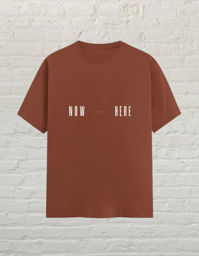 CoreTeez unisex copper cotton t-shirt featuring a minimalist white Now Here graphic print against a brick wall.