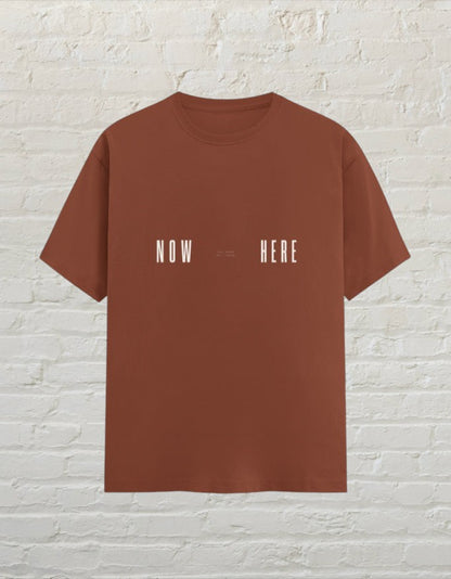 CoreTeez unisex copper cotton t-shirt featuring a minimalist white Now Here graphic print against a brick wall.