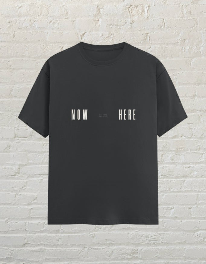 CoreTeez unisex steel grey cotton t-shirt featuring a minimalist Now Here graphic print against a white brick background.